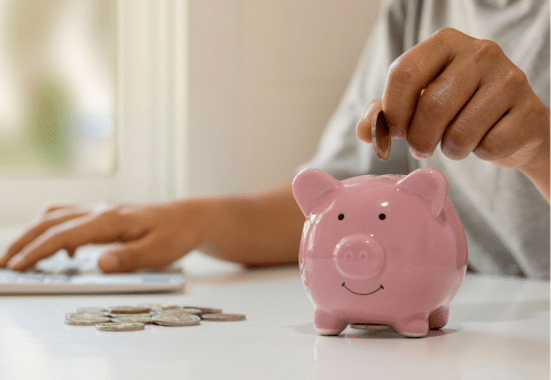 Main déposant une pièce dans une tirelire rose sur un bureau, symbolisant l’optimisation du patrimoine et la maîtrise des coûts liés à la transmission d’entreprise – accompagnement juridique et fiscal par un cabinet d’expertise comptable.

