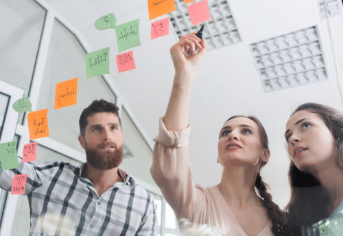 Trois jeunes professionnels collaborent devant une vitre couverte de post-it colorés, symbolisant le travail d’analyse et de visualisation des données comptables pour en extraire des indicateurs de gestion pertinents et guider les décisions stratégiques de l’entreprise.

