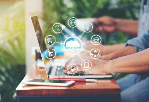 Personne travaillant sur un ordinateur portable en extérieur, avec une illustration en surimpression représentant la dématérialisation et l’automatisation des flux (cloud, données, achats, banque, RH, etc.), symbole de la transformation digitale des fonctions comptables.