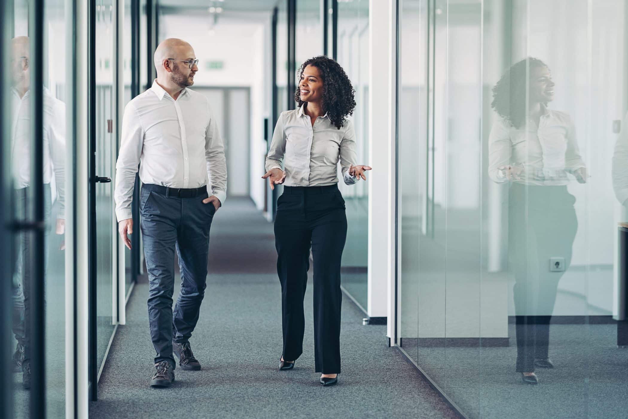 Deux professionnels échangent en marchant dans un couloir d’entreprise, incarnant la relation de proximité, d’écoute et de conseil qu’un expert-comptable peut entretenir avec ses clients pour les accompagner au quotidien dans leurs décisions stratégiques, comptables et fiscales.

