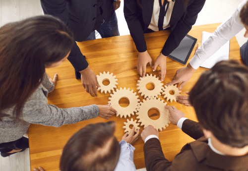 Plusieurs collaborateurs réunis autour d’une table assemblent des engrenages en bois. Illustration de la conduite du changement en entreprise, reposant sur une démarche collaborative, structurée et progressive pour faire fonctionner ensemble les différents leviers de transformation.

